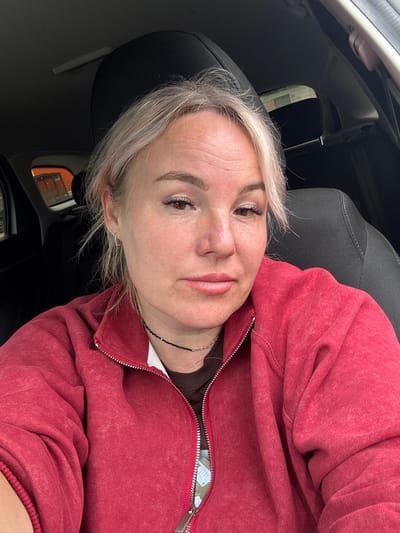 A woman with blonde hair and light skin, wearing a red zip-up hoodie over a dark shirt and a beaded necklace, is sitting in the driver's seat of a car. She is looking towards the camera with her mouth slightly open. The interior of the car, including a black headrest and seat, is visible. Outside the car window, some orange structures and a building can be seen. The image appears to have been taken during the daytime.