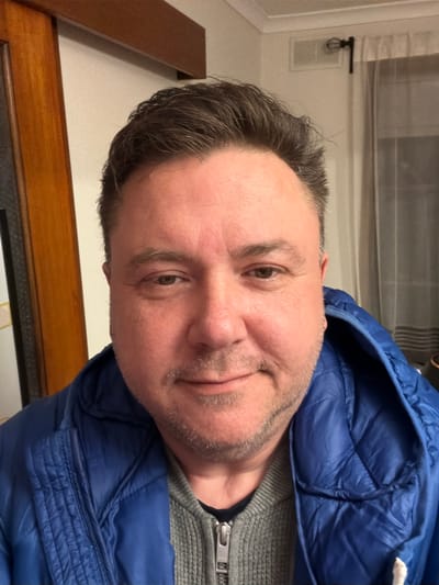 A middle-aged Caucasian man with short, dark brown hair and a light beard is pictured in a selfie taken indoors. He is wearing a blue puffer jacket over a grey zippered sweater. The background includes a wooden doorframe to the left, a white wall with a vent and a curtained window to the right. The lighting suggests an indoor setting at night, possibly in a residential area. The image does not contain specific identifiable landmarks or features of Parkdale, Australia.