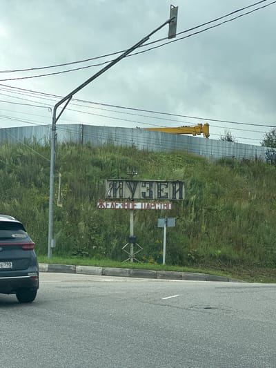 The image shows a street scene in Aristovo, Russia. A dark-colored Kia Sportage car is parked on the left side of the frame. In the background, a hillside covered in dense green grass and weeds rises up, topped by a corrugated metal fence. A large yellow construction crane is visible above the fence. Below the fence, a sign with Cyrillic lettering is mounted on a pole. The top line of text appears to read "МУЗЕИ" (Museum), and the bottom line appears to read "ЖЕЛЕЗНОЕ ЦАРСТВО" (Iron Kingdom). A street sign is also visible below the main sign. The sky is overcast and gray. The road in the foreground is asphalt, with a white line marking a lane.