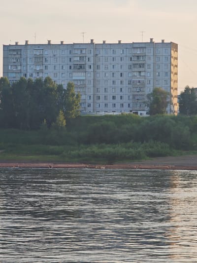 A multi-story apartment building stands on a grassy embankment overlooking a body of water. The building is light gray with numerous windows and balconies. Trees and vegetation cover the embankment. In the foreground, the water displays rippling reflections. A raft of logs is visible near the shore. The scene appears to be in the city of Lesosibirsk, Russia, given the architectural style of the building and the natural environment.