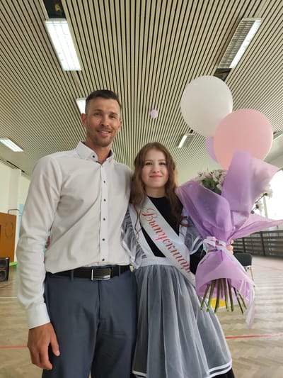 In an indoor setting, likely a school gymnasium or auditorium in Gremyachinsk, Russia, a man and a young woman stand together. The man, on the left, is wearing a white collared shirt, gray trousers with a black belt. The young woman, likely a graduate, is wearing a dark dress, a gray apron dress with white trim, and a sash with Cyrillic text on it. She holds a bouquet of flowers wrapped in lavender paper and three balloons (white, pink, and a smaller, unidentified color). The background includes a ceiling with horizontal stripes, lighting fixtures, and a wooden floor.