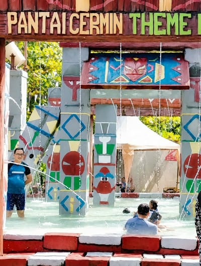 In Pantai Cermin Kiri, Indonesia, a group of people are enjoying a day at a theme park water attraction. In the foreground, a man wearing glasses and a blue and black t-shirt with blue shorts stands partially submerged in the water, smiling. Several other individuals are also in the water, including a person in a blue shirt and others partially visible. The water attraction features colorful, cartoonish pillars with various designs, including a red face with a stern expression and a creature with blue hair and a red body. Water sprays from the structure, creating a refreshing environment. Above the attraction, a wooden sign reads "PANTAI CERMIN THEME PARK" in large, colorful letters. The background shows trees and a tent structure. The scene captures a lively and fun atmosphere at the water park.