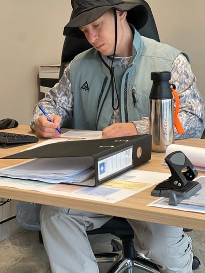 A person is sitting at a desk indoors. The individual is wearing a gray jacket, a hat, and gray pants. They are holding a blue pen and appear to be writing on a document in front of them. On the desk are a metal water bottle, a hole punch, a black binder filled with papers, and a keyboard. The scene appears to be an office setting. It is not possible to determine the location as Зеленогорское сельское поселение, Ukraine, from the given image.