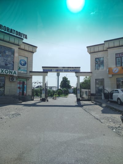The image depicts an entrance to what appears to be a medical complex or a collection of medical facilities in Shahrisabz City, Uzbekistan. The main structure is an archway with a prominent sign that reads "SHAHAR TIBBIYOT BIRLASHMASI," which translates to "City Medical Association." To the left of the archway, a building features a sign with Cyrillic text that says "LABORATORIYA," indicating a laboratory. Below this, another sign reads "APTEKA IDEAL," suggesting a pharmacy. On the right side of the archway, a building has a sign that begins with "TER" and also has some images and text in an unknown language. A white car with its driver's side door open is parked to the right of the entrance, partially obstructing the view of the building. The road in front of the entrance is paved, with some debris and gravel on the sides. Beyond the archway, a paved road leads further into the complex, lined with trees and smaller buildings. The sky is bright, with a strong sun creating a lens flare effect at the top of the image. The overall impression is of a functional, institutional entrance in a moderately urban or suburban setting.