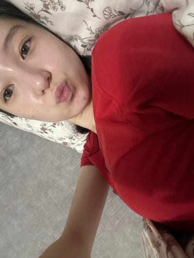 A young woman with fair skin and dark hair is lying on a bed with a floral patterned pillow. She is wearing a red t-shirt and is puckering her lips. Her eyes are open and looking towards the camera. The background consists of a textured gray wall or floor. The image is taken from a high angle, looking down at the woman. There is no indication of the city of Nookat, Kyrgyzstan.