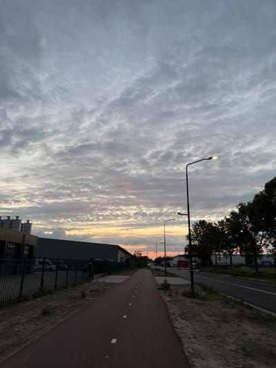 This image depicts a path next to a road with streetlights, bordered by industrial buildings and trees, under a cloudy sky that shows hints of sunset colors. The location is identified as Boxtel, Netherlands. The scene appears to be an urban or suburban area with infrastructure supporting transportation and industry. The path is a reddish-brown color, possibly a bike path or running track, with white markings. A black metal fence runs along the left side of the path, separating it from a grey building with cylindrical silos. On the right, a road with white lane markings is visible, with several streetlights lining it. A row of dark green trees stands between the road and further industrial structures in the background. The sky is filled with textured grey and white clouds, with soft pink and orange hues near the horizon, indicating either dawn or dusk. No people are visible in the frame.