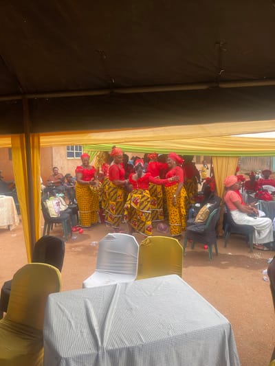 The image displays an outdoor social gathering taking place under a large, temporary canopy structure in Issele Uku, Nigeria. The ground consists of reddish-brown dirt. A prominent group of approximately eight women stands in the central frame, all dressed in identical attire: red lace blouses, yellow patterned ankle-length skirts, and matching red headwraps (gele). One woman within this group holds a patterned, woven basket-like object. Another woman in this group is observed bending slightly, engaging with a shorter individual who is also wearing similar patterned attire.
To the left of the central group, a woman in a black and brown patterned top and black trousers is seated at a white-clothed table. To the far right, a woman wearing a light orange top and a white and black striped skirt is seated. Other individuals are seated further back, some also in red attire.
The overhead canopy is black, supported by poles, and internally draped with yellow and light green fabric panels. In the foreground, a table covered with a white and grey checkered tablecloth is present. Several chairs are visible, some covered with black, white, or yellow fabric covers. A light brown building with a white-framed window is visible in the background. Scattered small red items and clear plastic bottles/cups are on the ground.