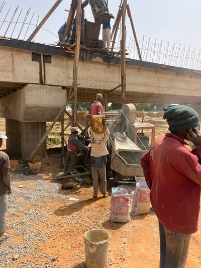 The image depicts an active bridge construction site in Wuciciri, Nigeria, under a clear, bright sky. An elevated concrete structure, supported by concrete pillars, is undergoing further construction. Rebar protrudes from the top surface of the structure, where concrete is being poured. Wooden scaffolding and a pulley system facilitate material transfer to the upper deck.
On the upper level of the structure, at least three male workers are present. One worker, dressed in a blue shirt and white pants, is guiding a large bucket of wet concrete as it is lowered onto the deck. Another worker in a light-colored shirt is also positioned on the structure.
On the reddish-brown, gravel-covered ground below, a grey motorized concrete mixer is active. One male worker, wearing a light-colored shirt and dark pants, carries a yellow bucket of material on his head, standing near the mixer. Another male worker in a red shirt operates the concrete mixer. Two white bags of "DANGOTE 3X" cement are visible on the ground beside the mixer. In the immediate foreground, a male worker in a red shirt and green knitted cap stands with hands on hips, speaking on a mobile phone. A grey bucket rests on the ground nearby. Distant green vegetation is visible in the background, beyond the construction area.