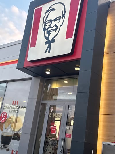 The image displays the exterior of a KFC restaurant in Belozem, Bulgaria. A large, prominent sign featuring the black outline of Colonel Sanders on a white background with red vertical stripes is mounted on the upper dark gray and red portion of the building facade. Below the sign, a glass double-door entrance with a dark gray frame is visible. To the left of the entrance, a large window displays a promotional advertisement for a "Donut with Bavarian Cream and Espresso," listing prices as 4.90 BGN (Bulgarian Leva) and 2.51 EUR. A smaller KFC logo is also visible on this window. The right-hand door features a red sticker with the text 'АРЪНИ / PULL' and a red rectangular sticker above it that reads 'РАБОТНО ВРЕМЕ' (Working Hours). The building's exterior incorporates dark gray composite panels and sections of light-toned wood paneling on the far right. Multiple circular, ceiling-mounted lights illuminate the entrance area. The upper left portion of the image shows a pale blue sky with scattered white clouds.