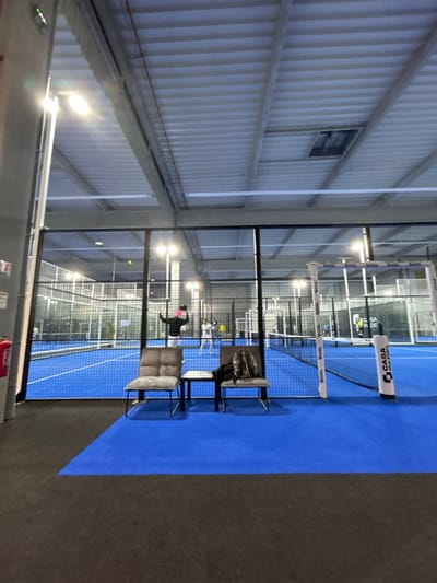 An indoor padel court, illuminated by overhead lights, displays blue playing surfaces enclosed by wire mesh walls. Two chairs sit adjacent to the court, near the black flooring. Players are visible on the court. The building features corrugated metal walls and a small window. A net bisects the court.
