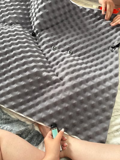 The image shows a gray foam mattress topper with an egg crate pattern.  A person on the right is holding a vape pen.  Another person's hands are visible on the left edge, apparently adjusting the topper.  Three metal latches are visible on the topper, seemingly part of a folding mechanism. The setting is indoors. It is not possible to verify any link to Boldino, Russia from the image.