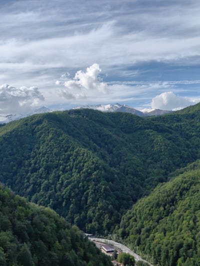 This vertical panoramic image presents a majestic mountain landscape in Эстосадок, Russia, showcasing a vibrant interplay of forested slopes, snow-capped peaks, and a dynamic sky. The scene is dominated by a succession of deeply verdant mountains and hills, densely covered with coniferous and deciduous trees, appearing in various shades of green depending on light and shadow.
In the lower midground, a prominent valley or gorge unfolds, through which a light-colored river winds its way. Along the riverbank, a small, isolated building with white walls and a dark roof is visible, suggesting a remote dwelling or lodge nestled within the natural environment. Further up on the forested slopes, two tall, metallic structures, likely power line towers or components of a ski lift system, punctuate the dense tree cover.
Beyond these forested peaks, in the background, a striking range of snow-capped mountains stretches across the horizon. Their rugged peaks, partially covered in white snow, stand out against the blue sky. The sky itself is a notable feature, displaying a mix of wispy, high-altitude cirrus clouds and more voluminous, fluffy cumulus clouds, all set against a clear blue backdrop.
The image conveys a sense of grandeur and tranquility, captured during what appears to be a bright day, likely late morning or early afternoon, with excellent visibility. The overall atmosphere is one of serene natural beauty, characteristic of a high-altitude mountain region. No discernible human activity or visible text is present within the frame.