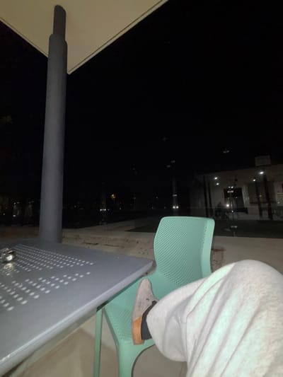 At night, a person wearing grey sweatpants sits on a light green perforated chair, with one foot resting on a grey metal surface. The scene looks out onto a cityscape, with lights scattered in the darkness. A metal pole runs alongside a structure, possibly part of the chair's design or a separate fixture. The perspective is tilted, adding a sense of height and overlooking the city below.