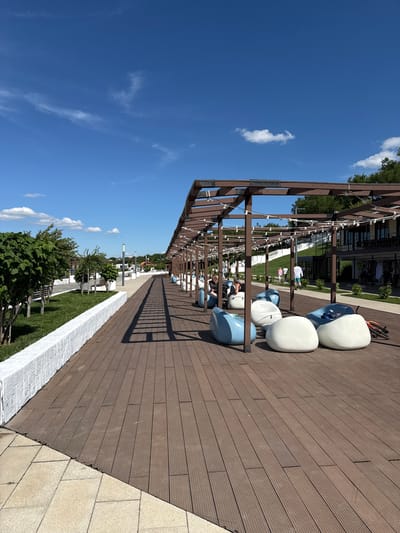 The image captures a vibrant outdoor public space on a bright, sunny day, likely in Taman, Russia. The scene features a long, modern wooden pergola structure, equipped with string lights, stretching from the foreground into the distance on the left side. Underneath and alongside this pergola, a spacious brown slatted wooden deck occupies the right side of the frame.

Arranged on the wooden deck are several contemporary, egg-shaped lounge chairs in white and light blue, providing seating for visitors. A bicycle is partially visible near some of these chairs. The strong sunlight casts prominent, elongated shadows from the pergola onto the deck, indicating midday or early afternoon.

In the middle ground, a strip of neatly maintained green grass is bordered by a low, white textured wall. This area is interspersed with small trees, bushes, and modern light poles. Further in the background, several people can be seen, some standing or walking on paved paths, while others are seated and relaxing. Modern buildings with large windows are visible beyond the landscaped area, suggesting a contemporary urban or resort setting.

The sky overhead is a clear, deep blue with a few scattered, wispy white clouds, indicating excellent weather conditions. The overall atmosphere conveyed is one of leisure and relaxation in a well-designed, modern outdoor environment. No visible text is present in the image.