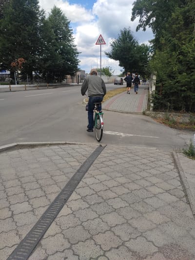 The image shows a man riding a bicycle on a street in Kostopil, Ukraine. The man is wearing a gray jacket and blue jeans. He is riding a green bicycle with a white fender. The street is paved with asphalt and has a white line in the middle. There are trees and buildings on the side of the street. There is also a pedestrian sidewalk with people walking on it. A traffic sign with a pedestrian symbol is visible in the background. The sky is cloudy.