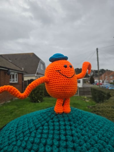 The scene captures a crocheted, spherical orange character with black button eyes and a stitched smile, resembling Mr. Tickle, positioned centrally on a textured, convex dark teal crocheted surface. The character wears a small blue crocheted hat, possesses two long, wavy orange arms—one extended to the viewer's left and the other raised and bent to the viewer's right—and short orange legs. The setting is a residential area in Upton, United Kingdom, under an overcast, light grey sky. In the mid-ground, a house with a light brown tiled roof, light-colored walls, and a white-framed window is visible to the left. Green grass and hedges are present between the foreground and the structures. A dark utility pole with overhead wires stands in the mid-right. The distant background reveals a road with several parked vehicles, including a light blue car and a dark-colored vehicle, alongside additional residential buildings.