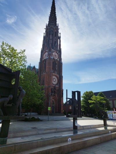 The image captures a view of the St. Mary's Church in Bremerhaven, Germany. The church features a tall, ornate brick spire with clock faces and a rose window. In the foreground, there are several bronze sculptures depicting figures, seemingly engaged in some form of labor or activity. These sculptures are part of a public art installation or memorial.

The scene appears to be set in an urban plaza or square, paved with stone and featuring tiered steps and water features, possibly a fountain area, although it is not actively flowing in this shot. Lush green trees provide a backdrop to the church and sculptures, indicating a pleasant outdoor setting.

The sky is bright blue with wispy white clouds, suggesting it is daytime, likely late morning or afternoon, with good weather. The sun is high, casting shadows and illuminating the scene.

To the right of the church, a modern building with a dark roof and multiple windows is visible. Further to the right, a vendor booth or kiosk with signage, including the word "HOTTER", is present, suggesting a public event or commercial activity in the vicinity.

The overall mood of the image is serene and somewhat contemplative, given the presence of the sculptures which could represent historical events or social commentary. The combination of the historic church architecture and contemporary public art creates a layered urban landscape. There is a green sign on a pole near the church entrance with some text, but it is too small to be legible.