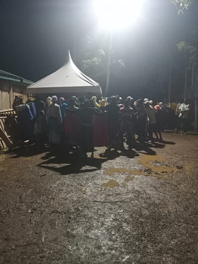 The image depicts a nighttime gathering in Butali Chegulo ward, Kenya. A bright artificial light source illuminates a group of approximately 20-30 individuals standing on muddy, wet ground with visible puddles and reflective surfaces. A white, conical-roofed tent occupies the center-left of the frame, under which some individuals are positioned. To the far left, a light-colored building with a green corrugated iron roof is partially visible, with stacked wooden materials against its wall. Trees are present in the dark background to the right. People are dressed in various casual attire, with some wearing head coverings. Their shadows are cast long across the muddy foreground. Individuals are mostly standing, some appearing to face each other or the tent. One person on the far right holds a stick or cane. No distinct actions or interactions are clearly discernible within the crowd; they appear to be congregated, possibly socializing or observing an event.
