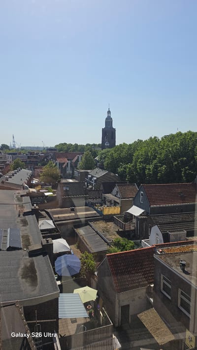 This media file depicts an aerial view of a European town, likely in Vlaardingen, Netherlands, taken during the daytime under clear skies. The scene showcases a dense urban landscape characterized by residential buildings with various roof types, including tiled and flat roofs. Lush green trees line the streets, adding a natural element to the built environment.
Prominently featured in the background is a tall, dark-colored church tower, a common architectural landmark in European towns. In the foreground, several residential rooftops are visible, some of which have small yards or patios. These areas are furnished with outdoor items such as umbrellas in shades of blue and yellow, and striped awnings, suggesting a pleasant, possibly residential, setting. One of the umbrellas is blue, another is a pale yellow, and a striped awning covers a patio area.
The image also captures glimpses of industrial elements in the distance, with what appear to be cranes and port structures hinting at a proximity to a harbor or industrial zone. The overall impression is one of a peaceful, sunny day in a developed urban area with a blend of historical and modern architecture. The only visible text is "Galaxy S26 Ultra" watermarked on the right side of the image.