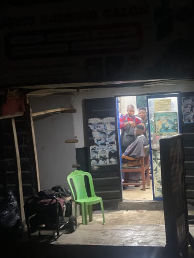 The image shows a dimly lit barbershop in what appears to be a low-income area, likely in Abraka, Nigeria. Through the doorway, a barber is cutting a customer's hair. Two other people are present in the shop. Outside the shop, there is a green plastic chair and a pushcart. The shop's sign, partially visible on the left, advertises "Dovis Barbering Salon," offering services such as "Hair Cut," "Tinjing & Rolling," "Home Service," "Clippers Maintenance," and "Dreadlocks & Diding." Contact phone numbers (09028344472, 09044768989) are listed. The address is "Beside Sambobn Hostel." The surrounding environment appears somewhat dilapidated, with unfinished construction materials visible in the foreground.