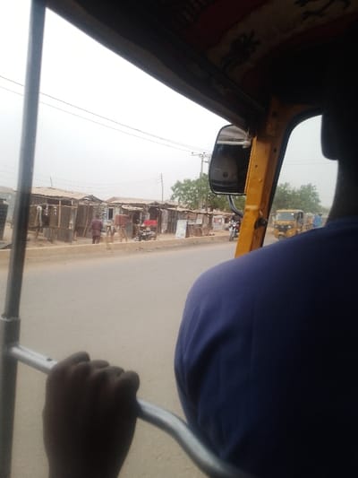 The image presents a street view in Lapai, Nigeria, observed from inside a moving yellow three-wheeled vehicle. In the immediate foreground, a dark-skinned hand grips a silver metal bar, positioned below the back of a person's head and shoulder wearing a dark blue shirt, consistent with a driver or front-seat passenger. The vehicle's yellow interior frame and a rearview mirror reflecting a light-colored surface are visible on the right.
Outside the vehicle, a paved two-lane road extends into the distance under a hazy, light grey sky. To the right of the road, a dirt median separates the asphalt from a row of approximately ten rustic market stalls constructed from wood and corrugated metal, each with a metal sheet roof. Several individuals are present on the dirt path in front of these stalls; one person in a maroon or dark red garment is walking away from the camera. A motorcycle is parked near the stalls. Further down the street, another yellow three-wheeled vehicle is discernible. Utility poles with overhead power lines are visible against the skyline, accompanied by sparse green trees in the background.