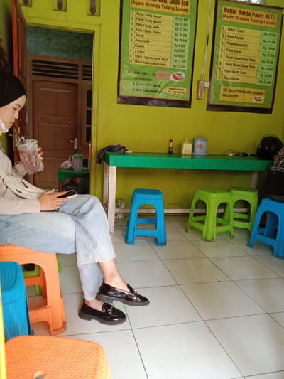 The image shows a woman seated indoors at what appears to be a casual eatery or waiting area, characterized by bright lime-green walls and a white tiled floor. The woman, positioned on the left, wears a black hijab, a white face mask pulled below her chin, a light-colored long-sleeved top, light blue wide-leg jeans, and black platform loafers with gold accents. She holds a transparent plastic cup with a dark beverage and a straw in her right hand, while her left hand is occupied with a pink smartphone.
Various plastic stools in orange, blue, and green are scattered on the floor. A long green table against the wall holds items such as a red condiment bottle, a yellow tissue dispenser, a light blue container, a black motorcycle helmet, and a dark cloth. A smaller green table near a wooden door and window in the background holds a clear bottle and a plastic bag.
Two price lists, written in Indonesian, are displayed on the green wall. The left menu, titled "Tanpa Nasi (Ayam Kremes Tulang Lunak)", lists individual items and prices (e.g., "Paha/Dada Hemat" for Rp 14.000, "Sayap" for Rp 10.500, "1 Ekor/Ingkung Ayam Jawa" for Rp 93.000), "Nasi Putih Rp 5000", "Pilihan Sambal", and a WhatsApp number (0859 5076 0440) for orders, featuring a "NUMANI Group" logo. The right menu, "Daftar Harga Paket AKTL (Ayam Kremes Tulang Lunak)", details package deals (e.g., "Paket 2 Ceker" for Rp 10.500, "Paket Kepala" for Rp 11.500, "Paket Bebek" for Rp 33.000) and notes that package prices include tea. The scene takes place during the daytime, and the woman appears engrossed in her phone, likely waiting or taking a break.