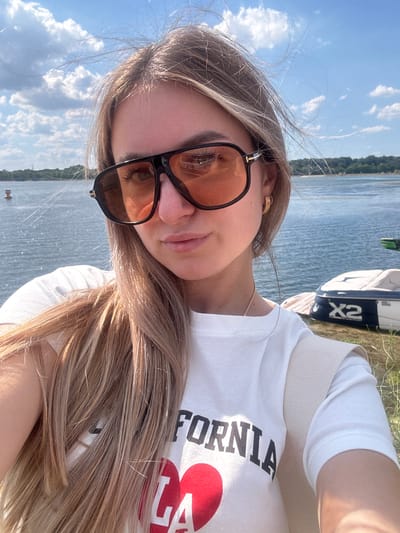 The media file is a selfie of a young woman outdoors. She has long, blonde hair and is wearing large, dark-rimmed sunglasses with orange lenses. She is also wearing a white t-shirt with black text that appears to say "CALIFORNIA" and a red heart graphic. A thin gold chain necklace is visible. Her hair is blowing in the wind, and her expression appears relaxed and content.
The background suggests a waterside location. The body of water appears to be a lake or river, with a tree-lined shore visible in the distance. There is a boat with "X2" written on its side docked on the shore behind her. The sky is blue with scattered white clouds, indicating a clear and sunny day. The lighting suggests it is daytime, likely late morning or afternoon. The overall impression is one of a casual, outdoor leisure activity, possibly near Altenheim, Germany, given the context. The woman's pose and the bright lighting contribute to a positive and cheerful mood.