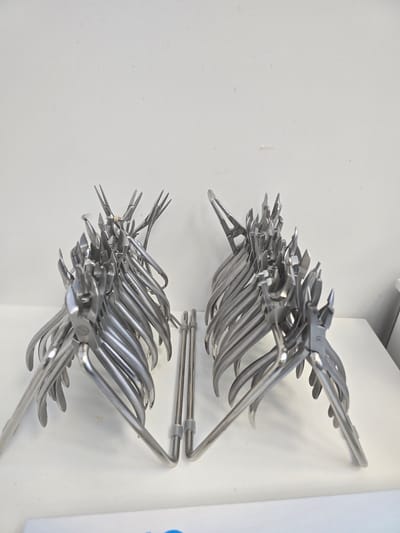 The image shows an arrangement of surgical instruments hanging on a white wall. The instruments appear to be stainless steel and of varying shapes and sizes, possibly for use in dental or orthodontic procedures. They are mounted on racks, creating two clusters on the wall. The location is Mouscron, Belgium.