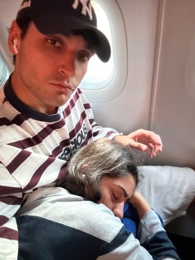 In this close-up, waist-up photograph taken inside an airplane cabin, a young man wearing a black New York Yankees baseball cap and a striped long-sleeved shirt with burgundy and white stripes is seen looking directly at the camera with a neutral expression. He has a white wireless earbud in his left ear. His right arm is resting on his lap, while his left arm is supporting another person.
The second person, also young, is asleep, leaning their head on the first person's arm. They have long, light brown hair and are wearing a blue and white striped long-sleeved shirt. Their eyes are closed, and their face is turned away from the camera. The background shows the interior of an airplane, with a window visible to the right, suggesting the location is on a flight. The presence of an airplane cabin indicates travel, and the question about Somma Lombardo, Italy suggests this flight might be arriving or departing from Milan Malpensa Airport (MXP), which is located in or near Somma Lombardo.