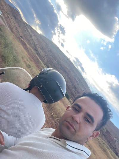 A low-angle selfie photograph shows two people on a motorcycle in a rural area, likely near Ait Mazigh, Morocco based on the landscape. The man taking the photo is fair-skinned, with dark hair and is wearing a white sweater with black trim. He is looking at the camera with a somewhat neutral expression. Behind him is a person wearing a black helmet and a white striped shirt, presumably the driver. The motorcycle has a transparent windscreen. The background features a hillside with sparse vegetation and a cloudy sky. The lighting appears to be from the late afternoon.