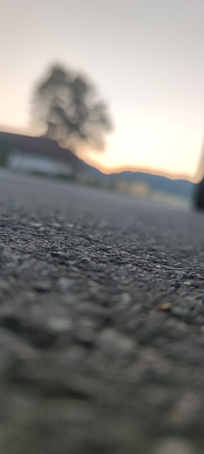 The image depicts a low-angle, close-up view of asphalt pavement, likely a road or path, with the texture of the surface in sharp focus. In the blurred background, a tree is visible against a bright, hazy sky that suggests either sunrise or sunset. Further in the distance, indistinct shapes of buildings and possibly hills are discernible. The overall impression is one of an outdoor scene, potentially in a semi-rural or suburban area, with the focus deliberately drawn to the granular detail of the ground. There are no people or animals visible in the image, and no specific actions or interactions are taking place. The scene is static and devoid of any distinguishing features that would allow for a precise geographical pinpointing within Liebenburg, Germany, beyond it being an outdoor paved area under a bright sky with some distant structures and vegetation.