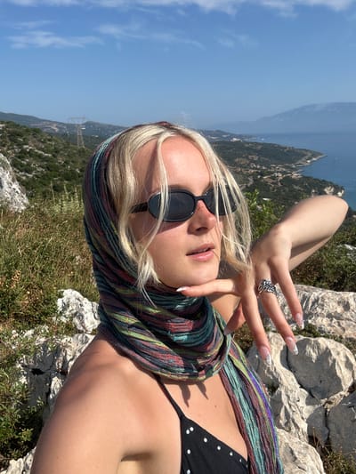 The media file shows a young woman with blonde hair and sunglasses taking a selfie on a rocky, grassy incline overlooking a body of water and a mountainous landscape. She is wearing a black bikini top and a colorful, striped scarf wrapped around her neck and shoulders. Her left hand is raised, adorned with long, decorated nails and a ring, and appears to be gesturing towards the camera. The setting suggests a sunny day with a clear blue sky. The background reveals a vast expanse of water, possibly a sea or ocean, with distant hills and mountains covered in greenery. The overall impression is one of leisure and enjoyment in a natural, scenic location. There is no visible text in the image.