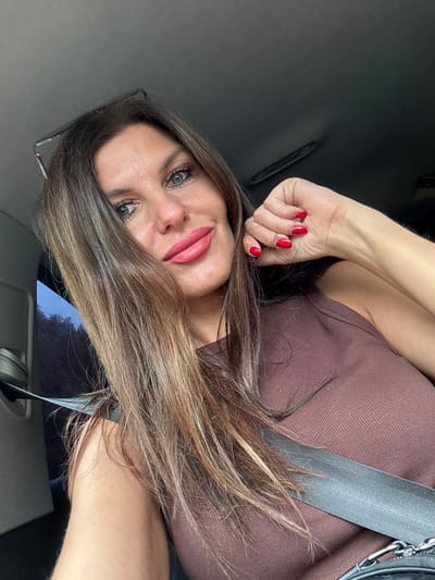A medium shot photograph shows a woman in the front seat of a vehicle, possibly a car. She has long, dark brown hair with lighter brown highlights, and it falls over her left shoulder. She is wearing a brown sleeveless top with a ribbed texture, and a grey seatbelt is fastened across her chest. Her left hand is raised towards her face, with bright red nail polish visible on her fingers. She is wearing makeup, including bold pink lipstick and mascara. Her eyes are open, and she is looking towards the camera. A pair of glasses rests on top of her head, partially obscured by her hair. The interior of the vehicle is dark grey, and a small portion of a dark-colored bag with a silver chain strap is visible in the lower right corner of the frame. The background outside the vehicle's window appears to be blurred and shows hints of trees and a slightly overcast sky. The image appears to be taken during daylight hours.