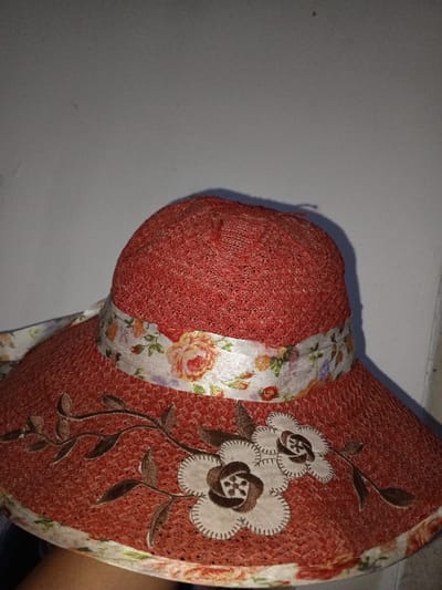 A reddish-orange woven hat with a wide brim is depicted in a close-up, positioned against a plain, light-grey wall. The hat features a white fabric band adorned with a multi-colored floral pattern, including orange, pink, and purple roses with green foliage, encircling the base of its crown. The brim of the hat is decorated with three off-white, multi-layered appliqué flowers with dark brown embroidered centers and outlines. These flowers are connected by dark brown embroidered stems and leaves. The underside edge of the brim also displays a continuation of the floral pattern from the band. No individuals or further environmental details are visible. This scene is identified as being in Na Sang, Laos.