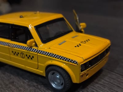 A close-up, high-angle, indoor shot features a yellow toy taxi with a checkered black and white stripe running along its side. The taxi has a single open door on the passenger side. A circular emblem with the letter 'T' is visible on the side of the taxi, near the front wheel. The taxi is placed on a dark, wooden surface. The background is dark and out of focus. The image does not contain any information about Novaya Usman', Russia.