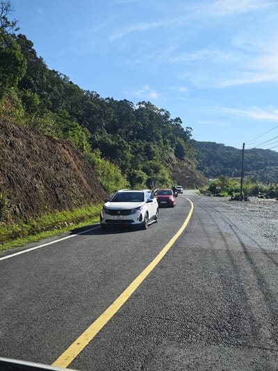 The image captures a winding, paved road with a distinct yellow line marking the center. Several vehicles are traveling on the road, including a white Peugeot SUV in the foreground, followed by a red sedan and two other cars further in the distance. The road is flanked by a steep, rocky embankment on the left, covered in sparse green vegetation. On the right, the terrain slopes upwards into a densely forested mountainside, with a clear blue sky dotted with wispy clouds overhead. A utility pole with visible wires stands on the right side of the road, near a gravelly patch. The scene appears to be a mountainous, rural area, likely a highway or a scenic route, with no visible signs of urban development in the immediate vicinity. The license plate on the white Peugeot reads "49A 493.87", suggesting a registration from Lam Dong province in Vietnam, which includes the city of Da Lat, but not specifically Nam Khanh Vinh which is a commune in the district of Nam Dan, Nghe An province. Without further contextual clues, identifying the precise location of Nam Khanh Vinh within this image is not possible.