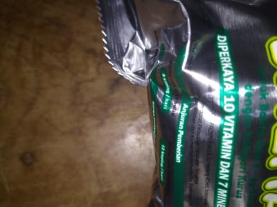 The image shows a close-up, oblique shot of a silver foil food package. Text is visible on the package, written in white and green lettering. Some of the text reads "DIPERKAYA 10 VITAMIN DAN 7 MINE" and "Anjuran Pemberian". The package is resting on a textured, brown surface that appears to be a wooden table. The lighting suggests the image was taken indoors, likely in a home or retail setting. No specific location in Gampongbaro, Indonesia can be determined from the image.
