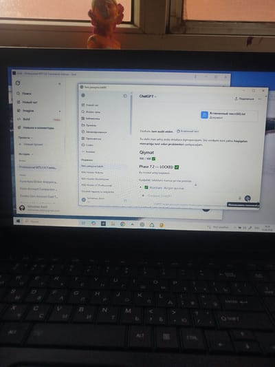 The image shows a close-up of a laptop screen displaying a computer program interface, likely a software development environment or a chat application. The screen is the primary subject, with a keyboard visible at the bottom, suggesting the photo was taken from the user's perspective.
The software interface shows a window with the title "Grok - Professional MT5 EA Framework Adessa - Grok" on the left and a "ChatGPT" window on the right. The ChatGPT window contains a conversation in what appears to be Azerbaijani, discussing an audit and phase completion. Text in Russian is also present, indicating potential multilingual use or system notifications. Notable details include a file name "Вставленный текст(60).txt" and a timestamp "16:20 30.06.2026" with "Hot weather" and "ENG" indicating the time, date, and language settings.
The overall scene is indoor, likely a home or office setting, with a blurred background suggesting an object or decoration above the laptop. The focus is clearly on the digital content displayed on the screen. The user's activity is implied to be interacting with the ChatGPT application, possibly for coding or technical assistance, given the software's name.