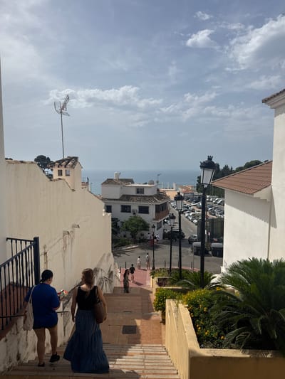 This is a vertical, eye-level, medium shot of a street and buildings in Benalmádena, Spain. The scene is viewed from a set of stairs descending towards a street, with buildings lining both sides of the stairs and the street. The sky is cloudy with patches of blue. In the foreground, two women are descending the stairs. The woman on the left wears a blue t-shirt and dark shorts, carrying a white tote bag. The woman on the right wears a black tank top and a long blue denim skirt, carrying a brown leather tote bag. Further down the stairs and on the street, other people can be seen walking. The street is lined with parked cars and lampposts. White buildings with terracotta tiled roofs are prominent throughout the scene. In the distance, the Mediterranean Sea is visible. Lush green plants and trees are scattered around the buildings and on the sides of the street. An antenna is visible atop one of the buildings.