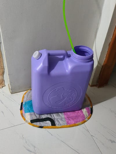A close-up shot shows a purple plastic water jug with a white cap and a green tube inserted into its opening. The jug sits on a colorful, quilted mat placed on a tiled floor. The jug has a handle on top and embossed text around a circular emblem that reads "H2O" and "100% VIRGIN MATERIAL". In the background, a plain gray wall meets a white trim and a wooden door frame. The lighting suggests an indoor setting, possibly during the day, with soft shadows cast by the objects. The overall scene appears to be in a residential or utility area within Cavite City, Philippines.