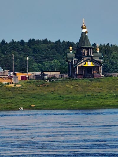 An Orthodox church with a green roof and golden domes stands on a grassy hill overlooking a body of water. The church is a wooden structure with a yellow emblem above the entrance. To the left of the church, there are several smaller wooden buildings and fences, suggesting a small village or settlement. The background is dominated by a dense forest of trees. The water in the foreground appears calm with visible ripples. The sky is a pale blue.