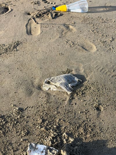 A close-up, eye-level shot of a sandy beach shows scattered debris and footprints. In the upper right corner, a clear plastic bottle with a yellow cap lies on its side. Slightly below and to the left of the bottle, a crumpled, translucent plastic bag is partially buried in the sand. The sand itself is coarse and textured, with small clumps and ripples. Several footprints, appearing to be from human feet, are visible in the sand, some deeply impressed and others fainter. In the bottom right and left corners, fragments of what appears to be a white object with dark markings are visible, partially obscured by sand. The lighting suggests it is either early morning or late afternoon, casting shadows on the sand. The scene is located on the beach of Pasi Rawa, Indonesia.