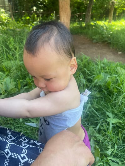 In Karakol, Kyrgyzstan, a young child is captured outdoors in a lush, green environment with trees and a dirt path visible in the background. The child, facing downwards and to the left, has dark hair and is wearing a light-colored, possibly white, wrap around their chest, with a glimpse of purple fabric underneath. The child's skin tone is light, and their eyelashes are dark and prominent. Their head is tilted, and their gaze is directed towards something out of frame. A person's arm and hand are visible in the foreground, partially obscuring the child's lower body and a blue and white patterned garment. The overall scene suggests a casual, outdoor moment.