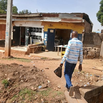 A man in a striped shirt and blue jeans walks away from the camera, carrying a black plastic bag in his right hand. He is walking on a dirt path in front of a small building with a corrugated metal roof. The building has a blue door and a small window. There is a concrete wall to the right of the building. The scene is located at coordinates 9.863 latitude and 8.867 longitude in the city of Kufang, Nigeria. The area is sparsely populated, with only a few other people visible in the distance. The ground is dusty and covered in dirt, and there is debris scattered around the path. The weather is sunny and dry. The building appears to be a small shop or stall, and there is some indication that there may be people inside, perhaps a customer and a shopkeeper. The man's expression is not visible. There are no other details available about the identity of the man or his purpose for walking away from the building.