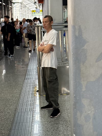 The image depicts a man standing on a platform, possibly a train or subway station. He is wearing a white t-shirt and dark pants, with his arms crossed. Several other people are visible in the background, also on the platform, suggesting a public transportation hub. The environment includes a tiled floor with a raised tactile paving strip for visually impaired individuals, metal railings, and a glass ceiling structure. A clock and directional signs are mounted on the wall in the distance. The overall scene is illuminated by artificial lighting and natural light filtering through the ceiling. The location is identified as Tiaodeng, China.