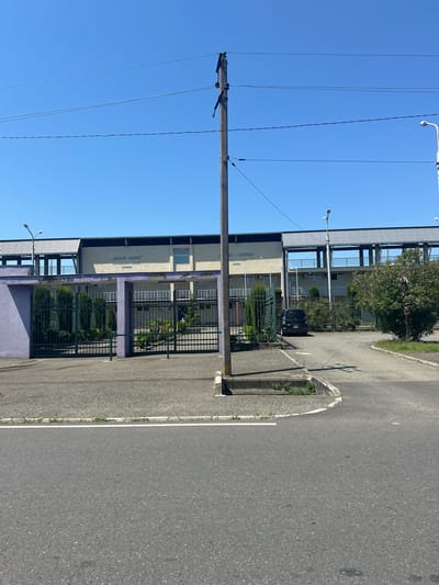 The image depicts the "Poti Arena" located in Poti, Georgia. In the foreground, there is an asphalt road curving to the right, with a concrete curb and a patch of grass along the edge. A concrete sidewalk or pedestrian path is also visible. A utility pole with wires runs vertically in the middle of the frame. Behind the utility pole, a dark gray fence with vertical bars encloses an area with manicured bushes and trees. To the left of the fence, a lavender-colored structure is visible. The main building of the Poti Arena is a long, light-colored structure with a ribbed metal roof extending over a covered walkway. The building has several windows and air conditioning units visible. Text in Georgian and English is present on the building, indicating "POTI ARENA" and other affiliations. A black SUV is parked inside the fenced area. The sky is clear and blue.