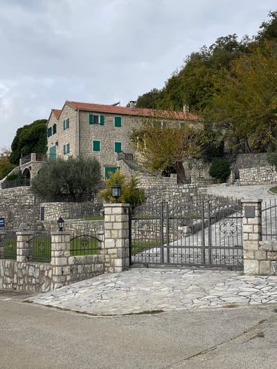 An exterior long shot captures a stone-built residence and its immediate surroundings in Đenjaši Česminovo, Montenegro, under an overcast sky. The multi-story building, constructed of light-colored rough-hewn stone blocks, features a red-tiled gabled roof with two visible chimneys. Numerous rectangular windows are fitted with closed green shutters, while one upper-story window on the main facade displays open green shutters. A dark-railed balcony is visible on the left side of the structure.

The property is situated on a terraced hillside, with extensive retaining walls and staircases built from the same light-colored stone, ascending towards and around the main dwelling. Mature trees, including a prominent olive tree to the left and several deciduous trees exhibiting autumnal foliage, are integrated into the landscaping. Denser green forest covers the higher slopes behind the house, particularly on the right.

In the foreground, a dark asphalt road runs horizontally. A substantial entrance gate, forged from dark metal with decorative scrollwork, blocks access to a driveway paved with irregular light-colored flagstones. This gate is flanked by two robust stone pillars, each capped with a black, lantern-style light fixture. A low stone wall with matching smaller pillars and dark metal fencing extends from these main pillars, defining the property's perimeter. A small, dark rectangular plaque is affixed to the right gate pillar. A partial blue sign is visible on a stone wall behind the left gate pillar. No individuals or active subjects are present in the scene.
