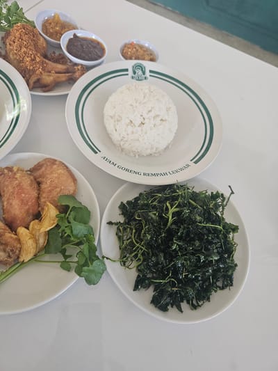 In Banyuurip, Indonesia, a table is set with a meal featuring fried chicken and rice. The scene is presented from a high-angle shot, focusing on a white table adorned with several plates of food. In the foreground, a white plate holds a generous portion of dark green, leafy vegetables, possibly stir-fried. To the left, another white plate contains pieces of fried chicken, some appearing coated in a crispy batter. Accompanying the chicken are several fried garlic chips and sprigs of green watercress. In the background, a plate of fluffy white rice sits on a larger plate that has a green and white striped rim and a logo with text that reads "AYAM GORENG REMPAH LEKWAR." Behind this, three small white bowls contain various dipping sauces or condiments. The overall setting suggests a casual dining experience, with the bright lighting indicating an indoor environment, possibly a restaurant. No people are visible in the image.