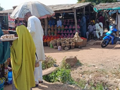 The image captures an outdoor market scene in Jere North, Nigeria. In the foreground-left, a person wearing a lime-green garment faces a partially visible individual holding a white tray containing approximately 15-20 square, light-colored food items. Adjacent to them, a man in a white tunic and a light-colored cap stands with his back to the viewer, looking towards the market stalls, under a large, light-colored market umbrella. The background features multiple rustic market stalls constructed with wooden frames and corrugated metal roofs. These stalls display various produce, including stacked red vegetables (likely tomatoes), light-colored root vegetables (likely onions or potatoes), and piles of garlic, arranged in baskets and sacks. Several men are visible within and around the stalls; three men are seated on a low bench or stools to the right of the central stall. A bright blue motorcycle is parked on the dirt path to the right of the stalls. The ground throughout the scene is unpaved dirt, with sparse green vegetation and a small drainage channel visible in the immediate foreground.