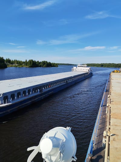 The image depicts a serene scene on a wide, dark blue river under a bright, clear blue sky dotted with scattered white clouds. The sun is high in the sky, indicating it is likely midday. The weather appears to be warm and pleasant.
The main subjects are two large barges navigating the river. The barge in the foreground, viewed from its deck, is a substantial vessel with a long, flat, light gray deck. Its blue hull is partially visible along the side. In the mid-ground, another barge is moving away from the camera, also with a long, flat, light gray deck. This barge has a white superstructure at its rear, which includes cabins and navigation equipment. The dark blue water of the river reflects the bright sky and the surrounding greenery.
The riverbanks are lined with dense, green trees, creating a lush natural backdrop. On the left bank, some houses and smaller structures are visible nestled among the trees, suggesting a populated area along the river. The barges are moving in the same direction, implying they are part of a convoy or traveling along the same route. The overall atmosphere is peaceful and tranquil, conveying a sense of calm movement and natural beauty. The location is identified as Nova, Russia, further specifying the setting. There is no visible text in the image.